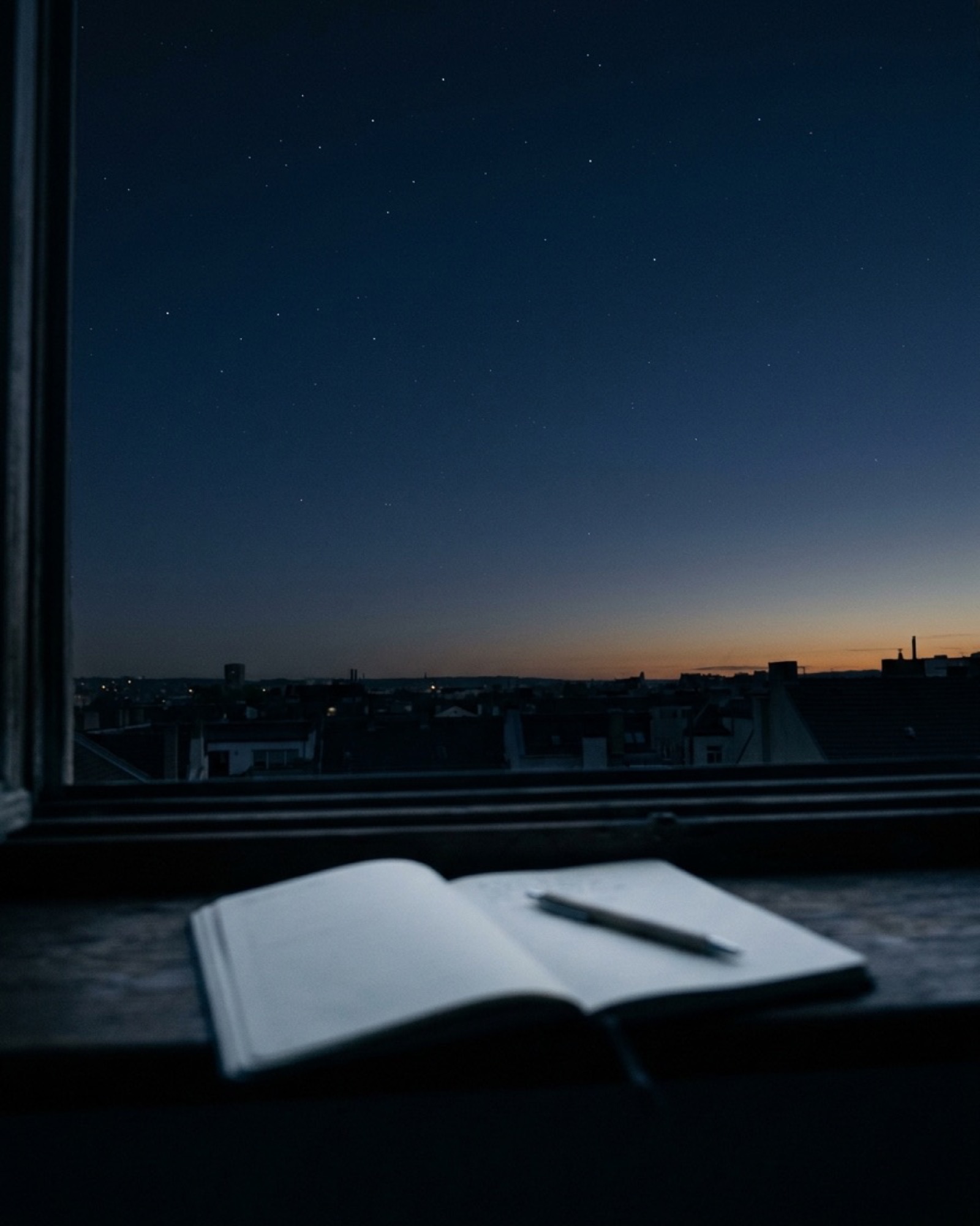 The same room at dawn seen from the window, a midnight sky over low rooftops and a notebook lying open on the sill.